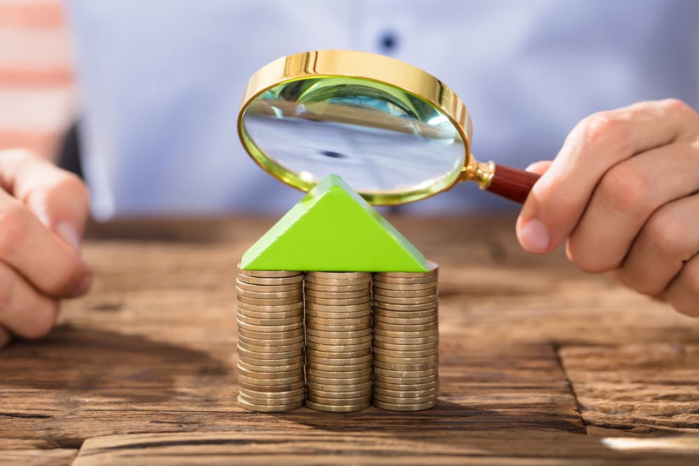 Close-up of a person holding a magnifying glass over a house made of coin stacks.