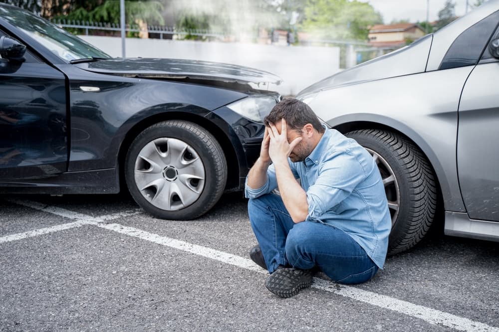 A distressed man sitting on the ground holding his head with two cars in an accident.