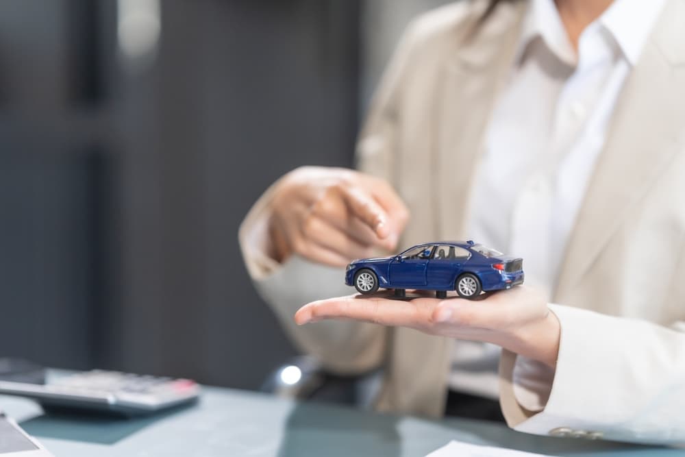 A person holding a small blue car model, pointing at it while sitting at a desk.