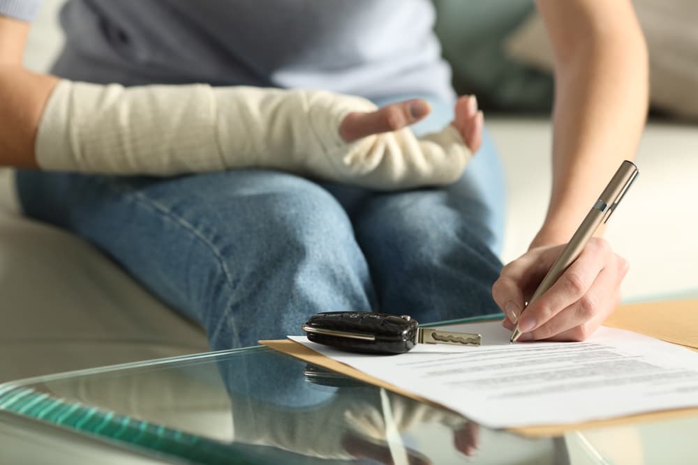 A person with a bandaged arm signing documents with car keys on the table nearby.