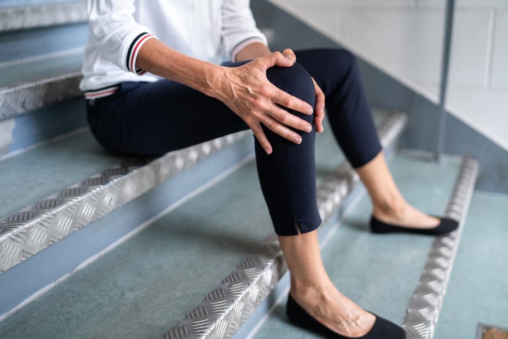 A person sits on stairs, holding their injured knee with one hand in pain.