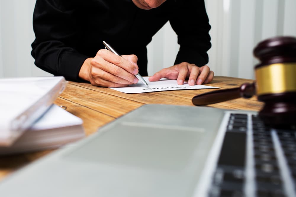 A person is signing a document at a desk with a gavel and laptop nearby.