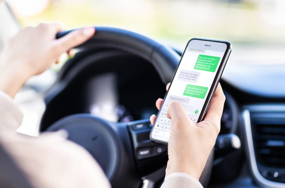 A person texting on a smartphone while driving, with one hand on the steering wheel.