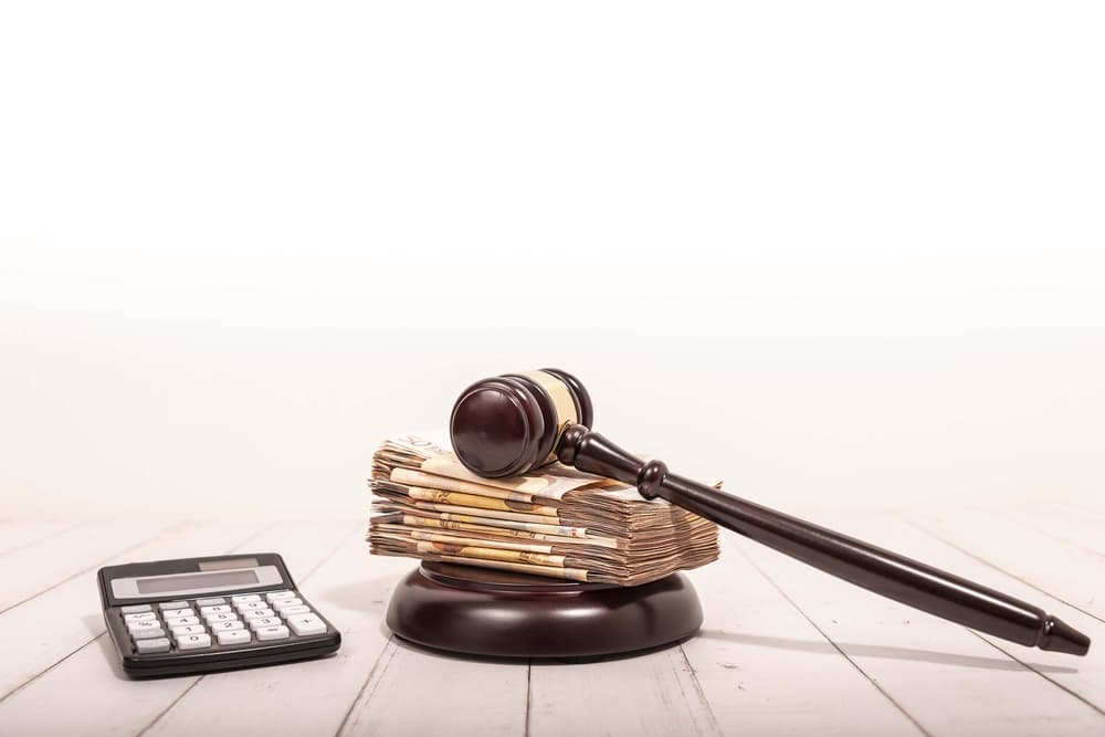 A judge's gavel rests on a stack of money next to a calculator on a table.