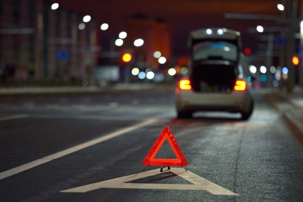 A red warning triangle on the road with a car in the background at night.