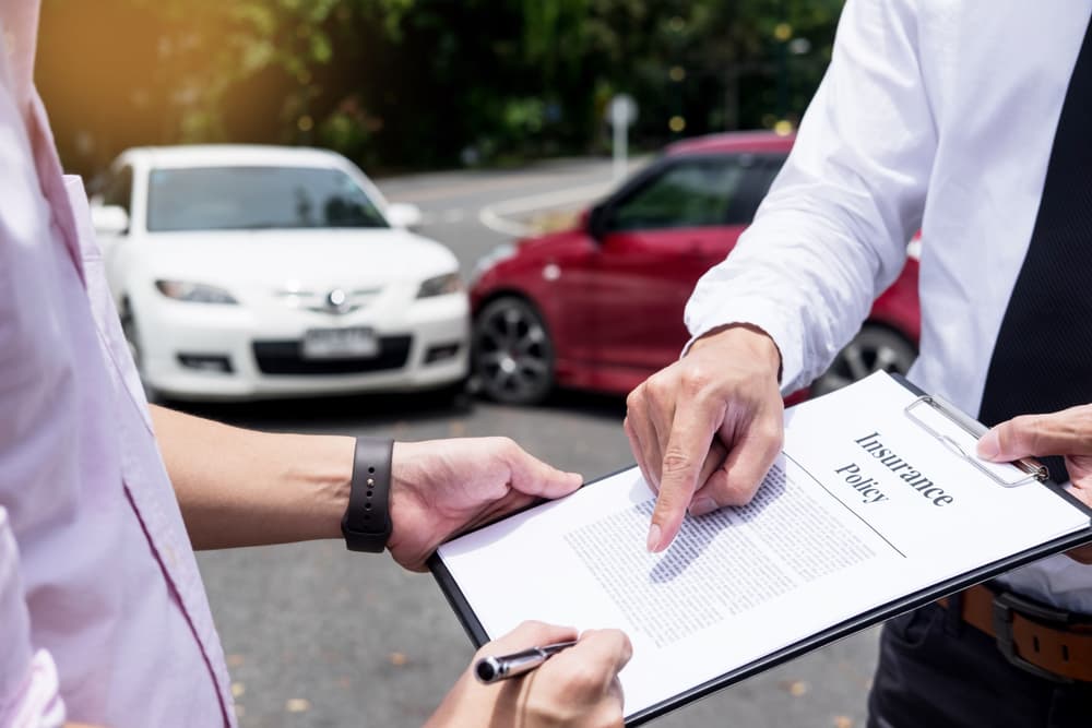 Two people discussing an insurance policy document with cars involved in an accident in the background.