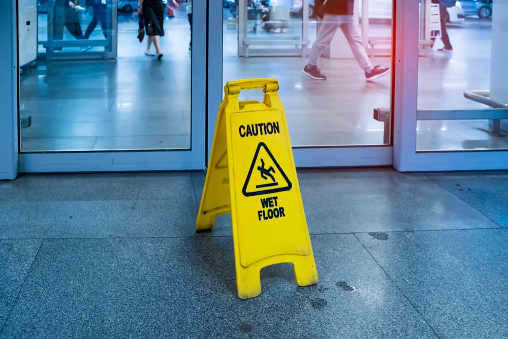 A yellow "Caution Wet Floor" sign stands in front of a glass entrance at a building.