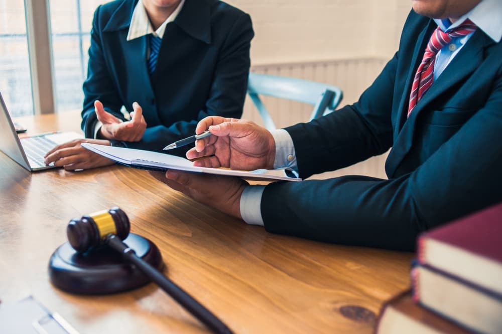 Two business professionals in suits discuss documents at a desk with a gavel nearby.