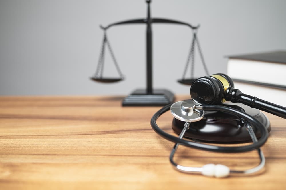 A gavel and stethoscope on a table with a justice scale and books in the background.