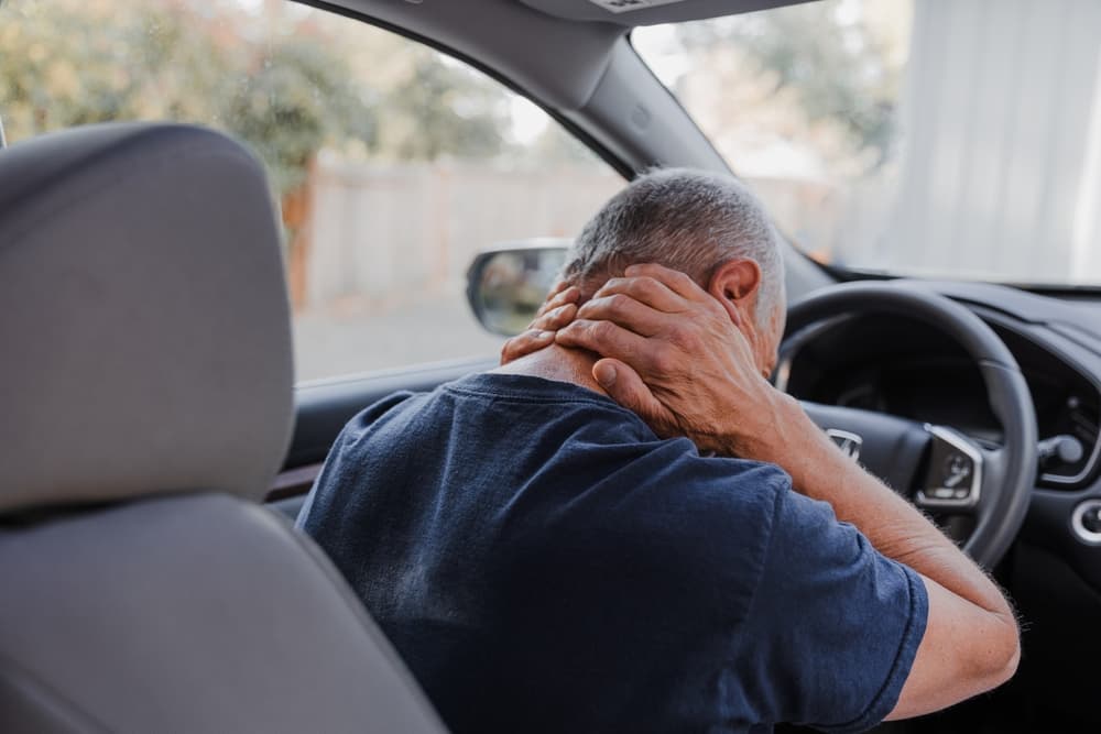 A man sits in a car, holding his neck with both hands, indicating whiplash pain.