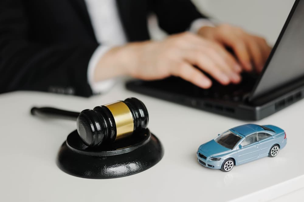 A gavel and toy car are placed on a desk, with a lawyer typing on a laptop.