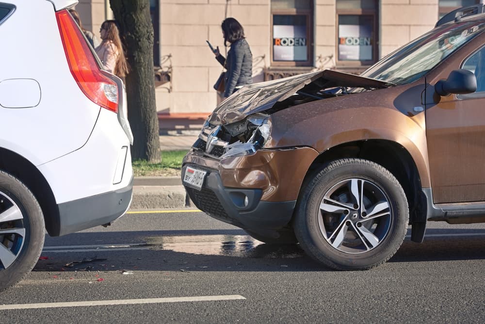 Two cars in a rear-end collision on the street with pedestrians nearby, inspecting damage.